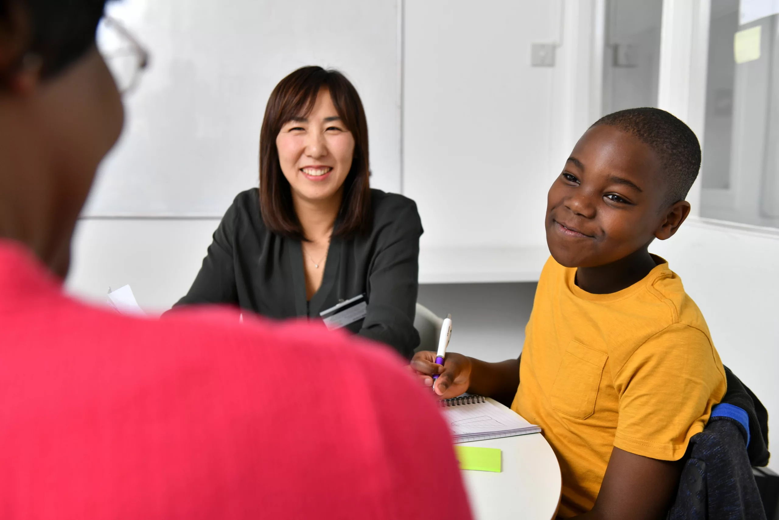 Two female professionals chat to a boy around a desk who is writing and smiling