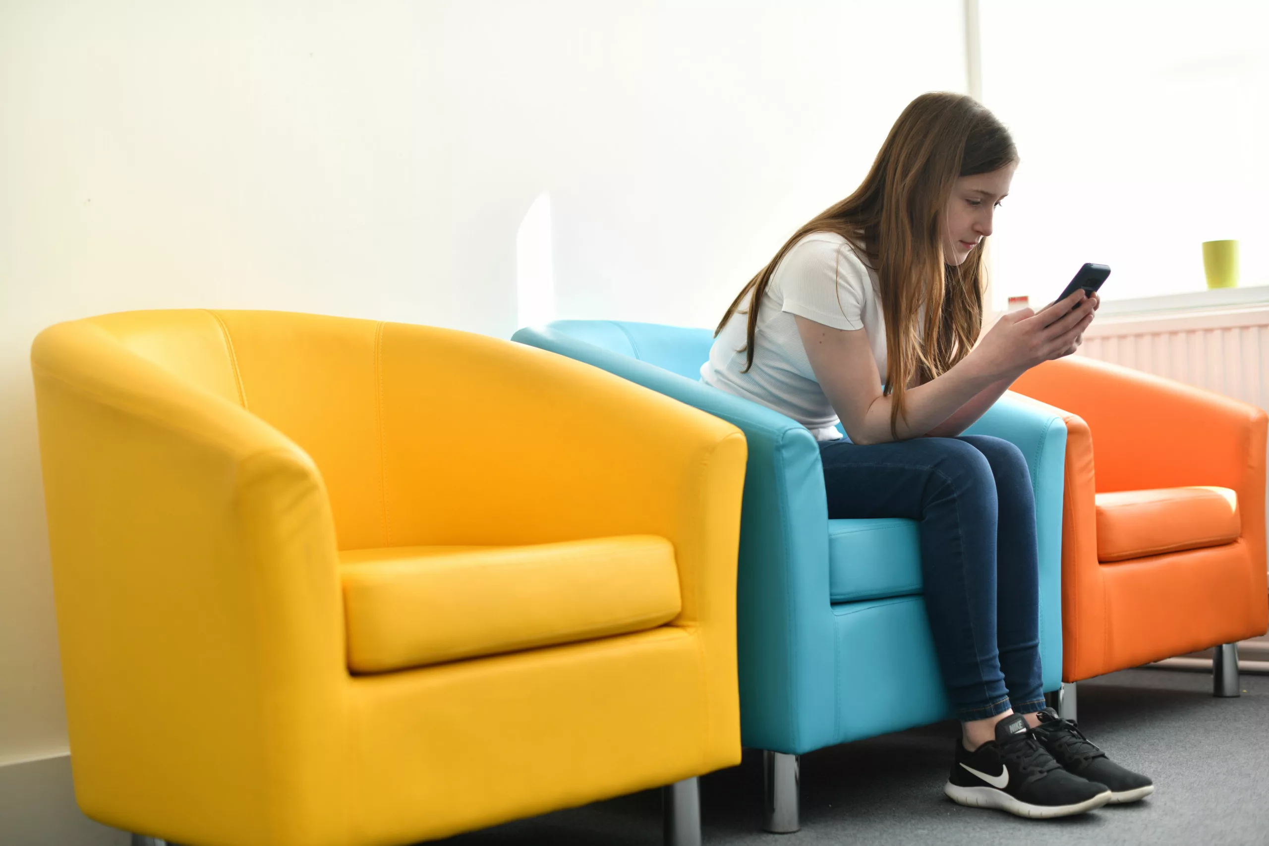 Teenage girl sits in the middle of three chairs looking at her mobile phone