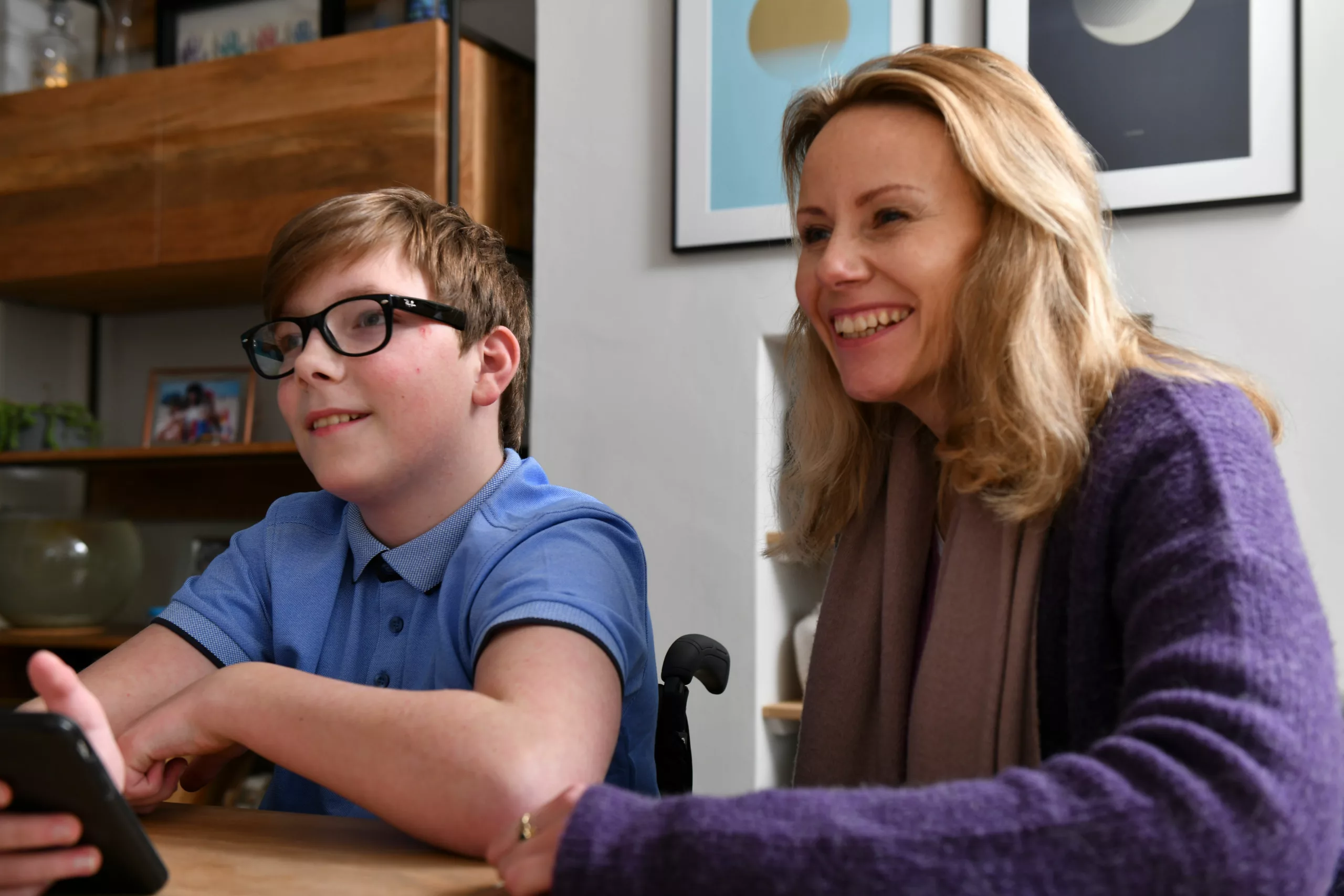 Boy sits alongside a woman at a table in a home holding his phone as they both smile at someone out of image