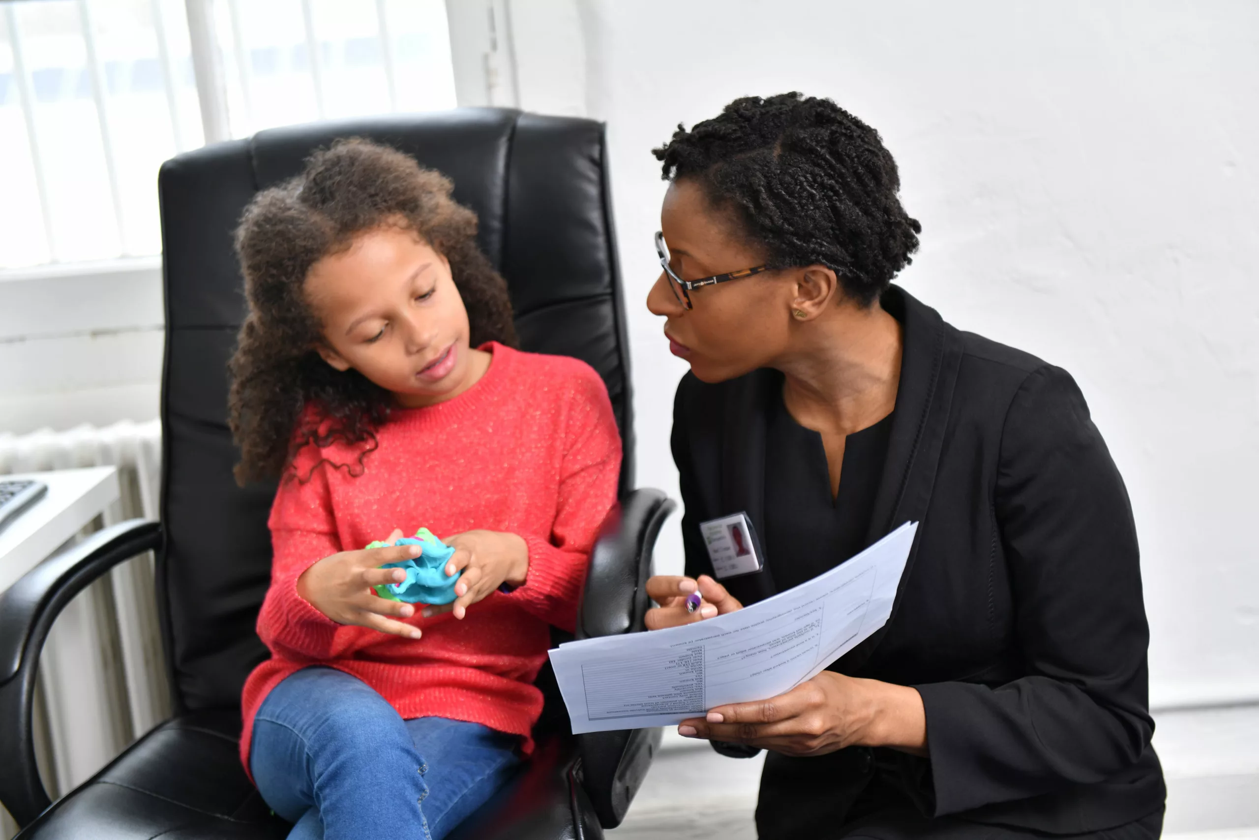 A young girl sat on a leather chair playing with a toy speaks to a female professional who is crouched alongside showing her a piece of paper