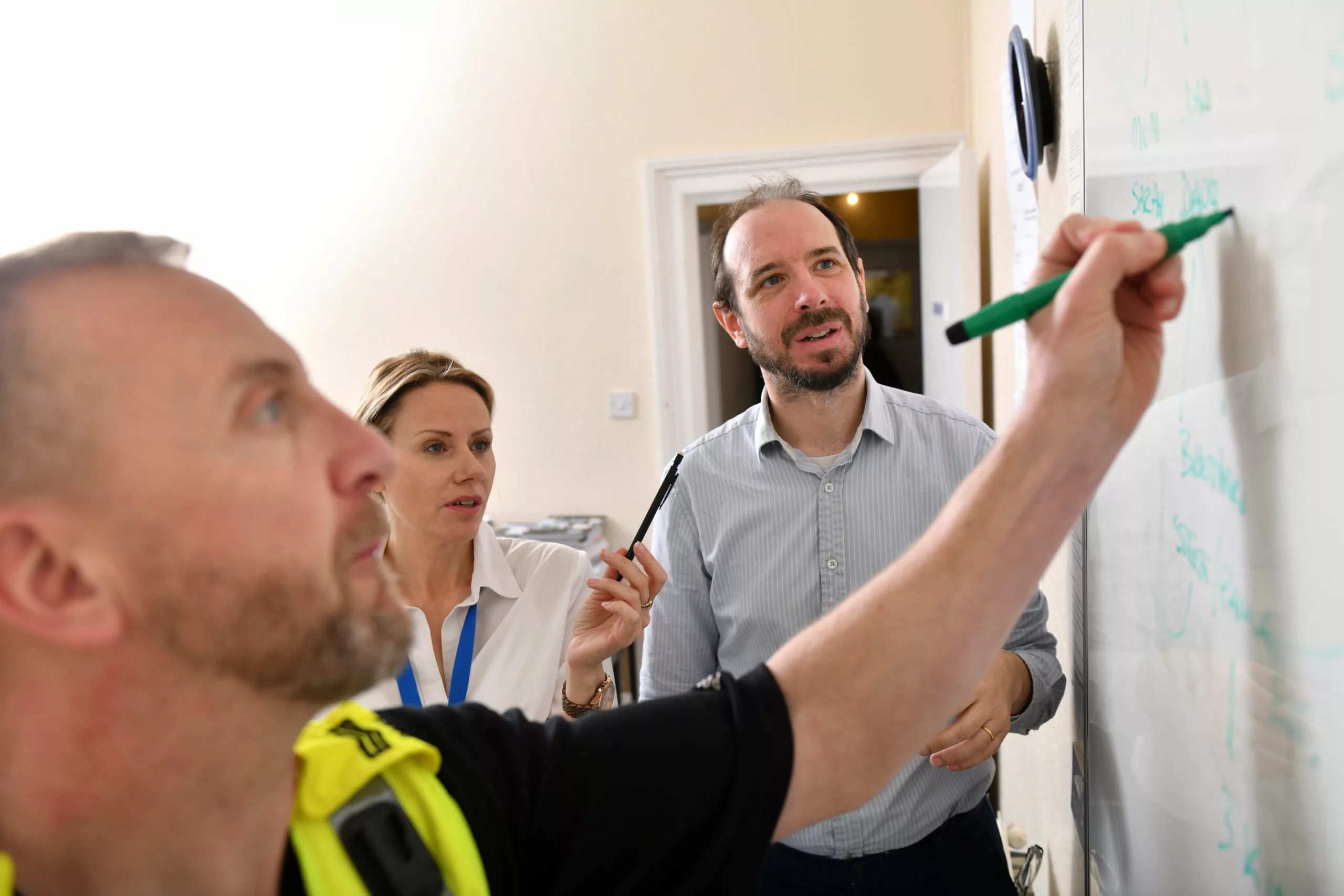 Three multi-agency professionals - one male, one female and another male in police uniform - write on a whiteboard