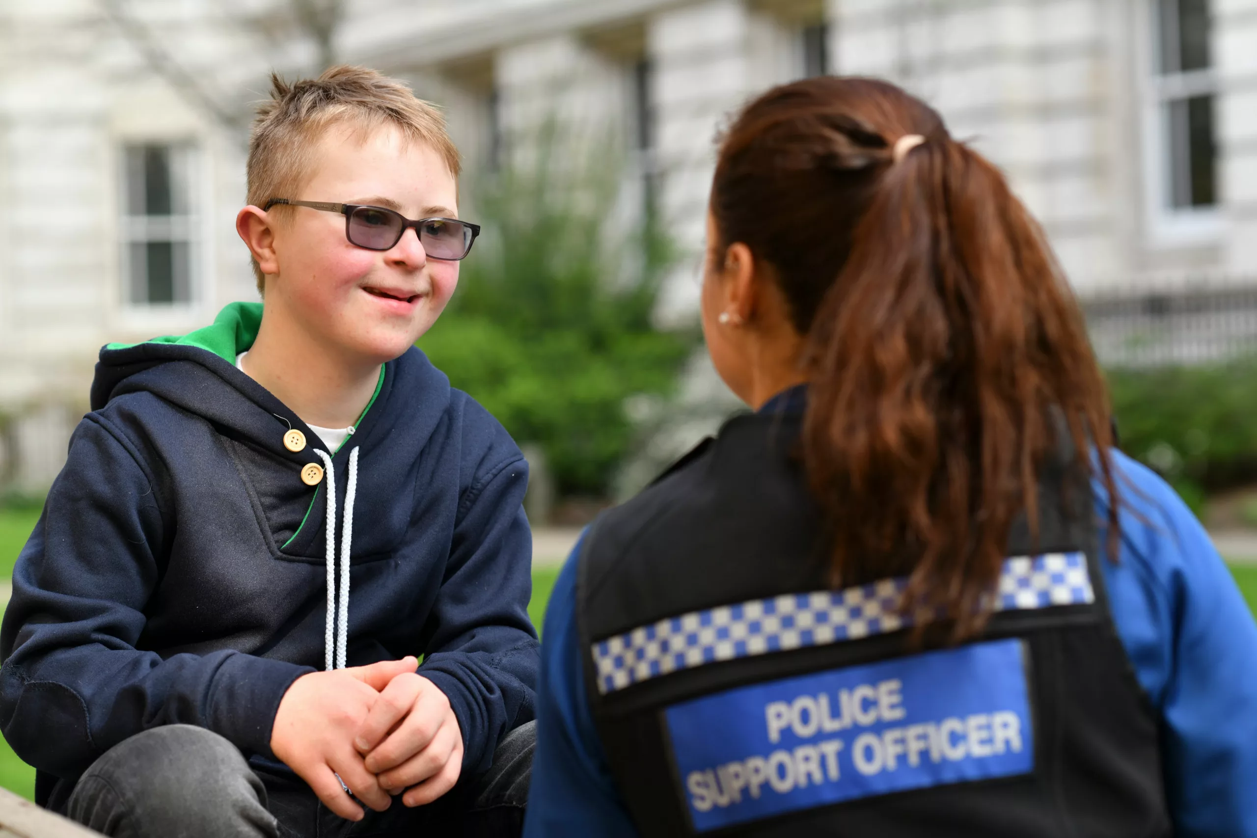 Young boy wearing shaded glasses speaks to a female police support officer outside and smiles