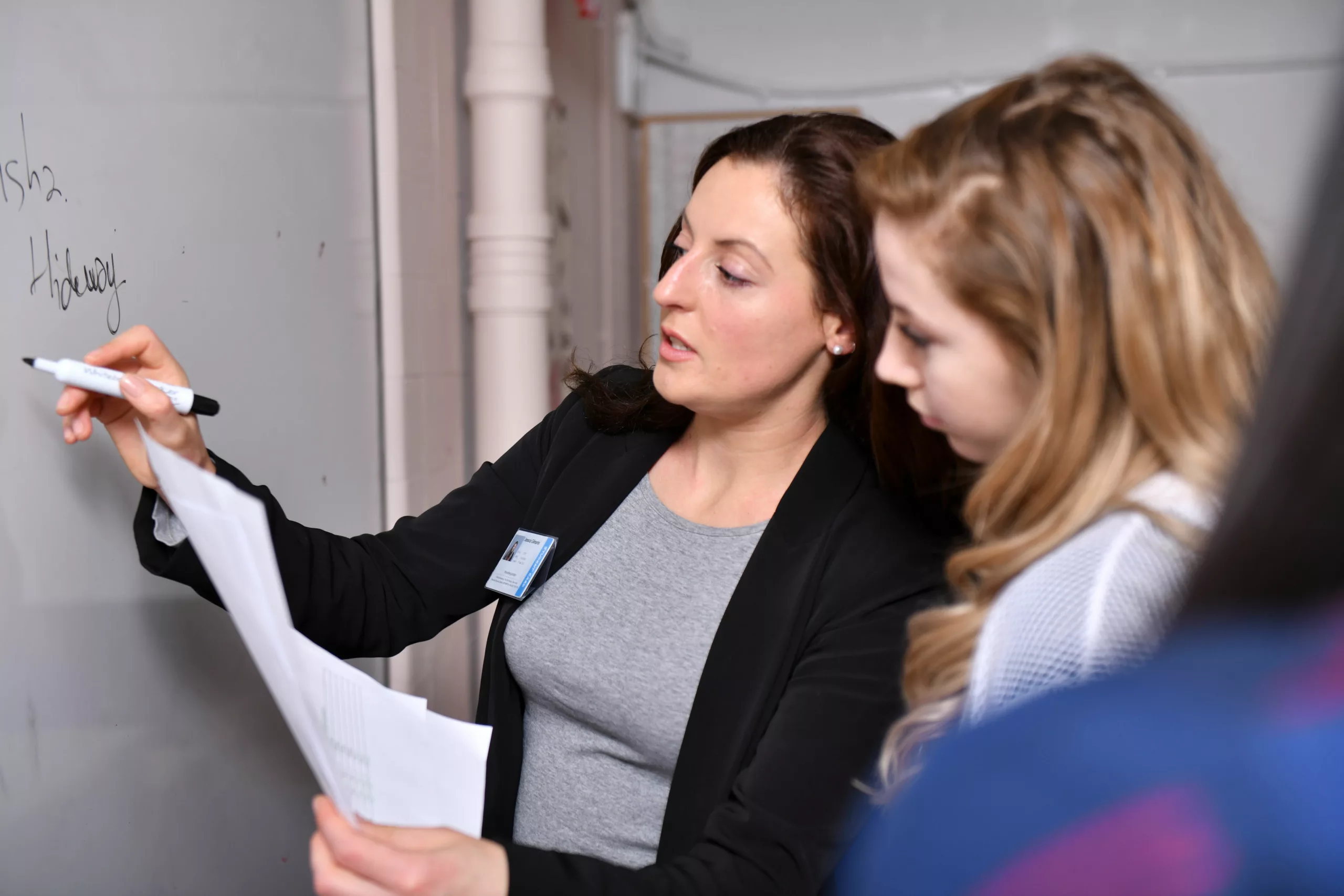 A female professional writes on a whiteboard whilst speaking to a teenage girl