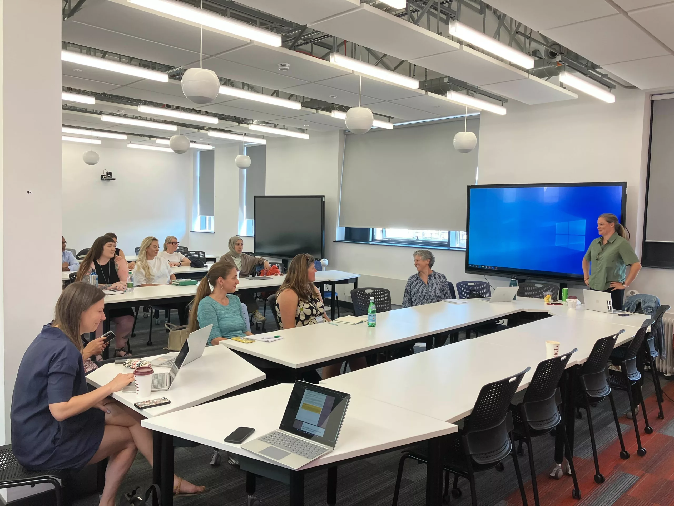 A group of female academics have a discussion in a workshop setting, with laptops and across two table