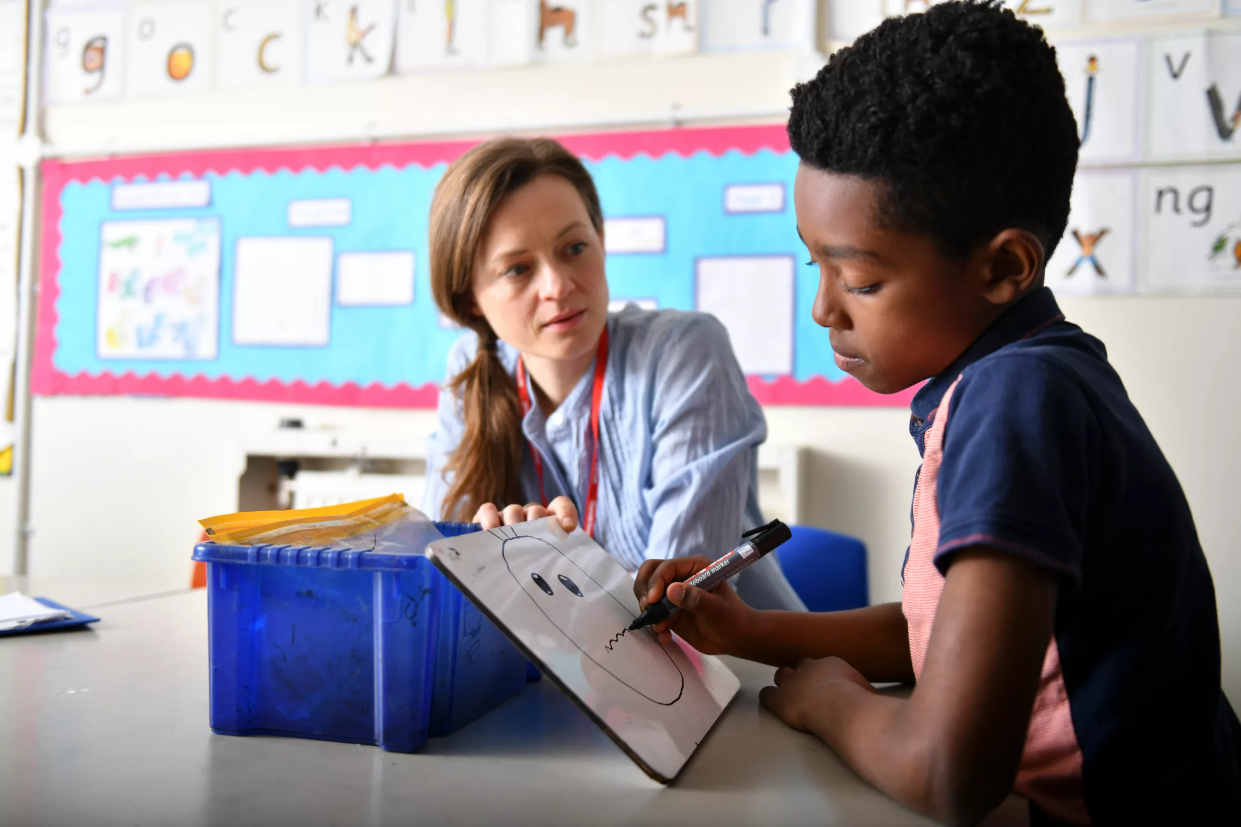 A female teacher sat at a table next to a boy in a classroom. The teacher looks concerned and is listening to the boy