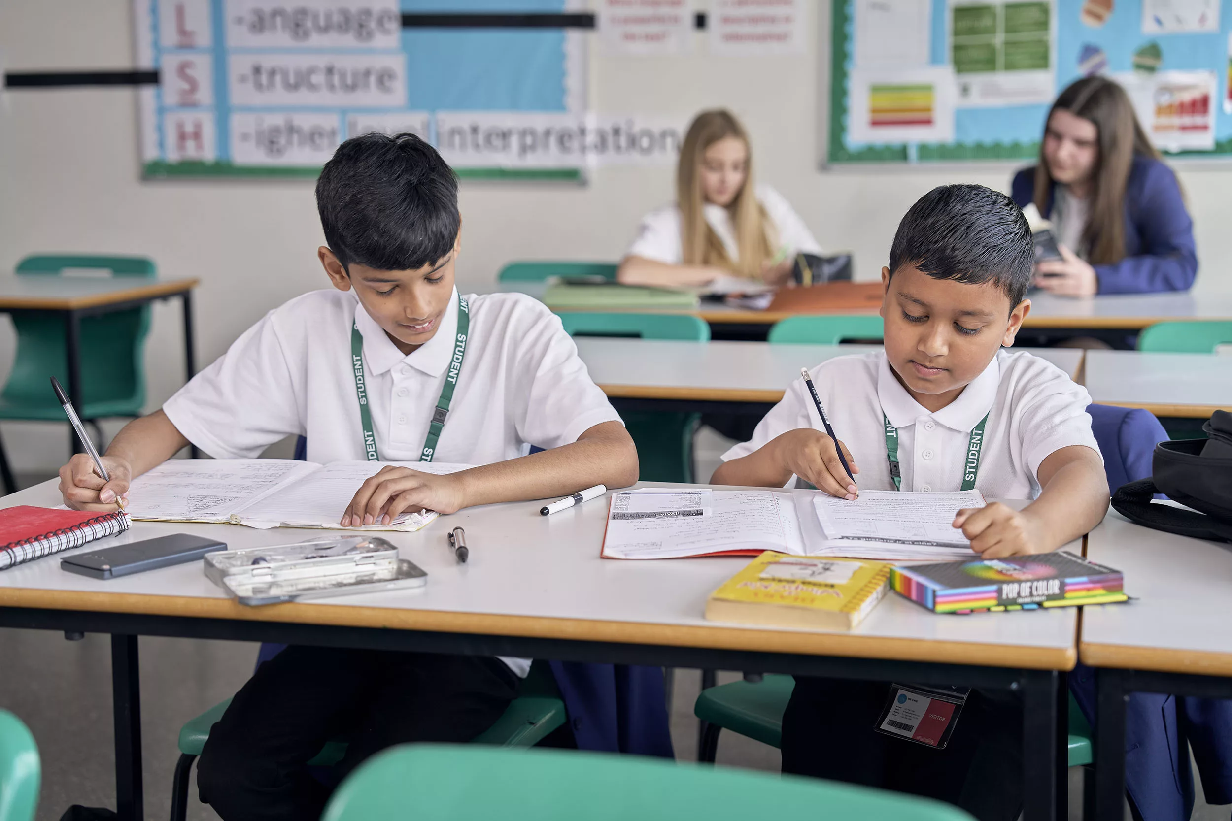 Two young boys sat behind a desk writing in a classroom, with two girls sat behind