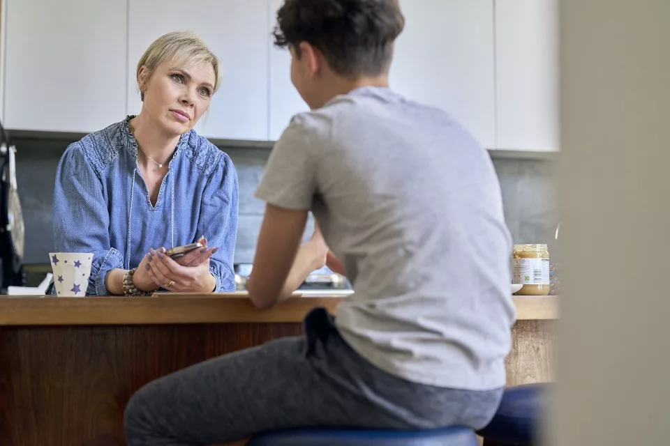 Mum holding phone looks across to speak to son at kitchen table