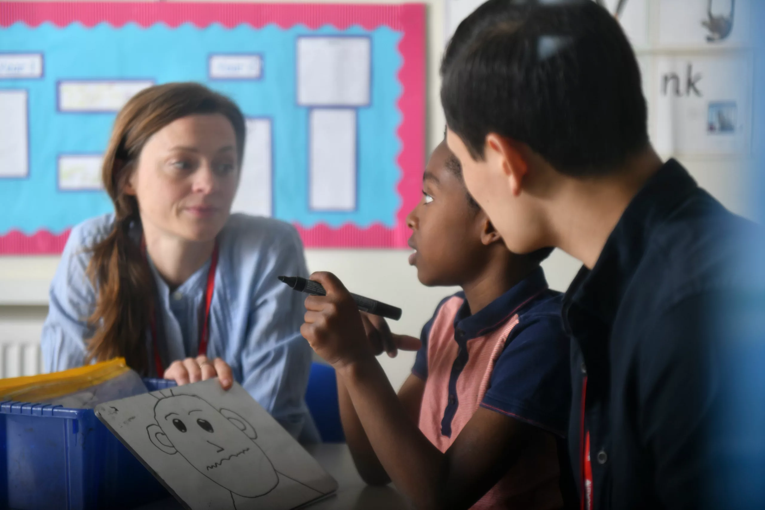 Two professionals speaking to a child in a classroom. The child is drawing on a small whiteboard with a drywipe pen.