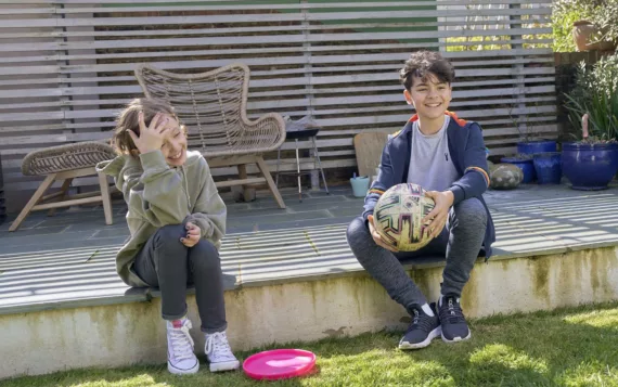 Girl with a frisbee at her feet and boy holding a football sit outside together in a garden. They're smiling at someone out of shot